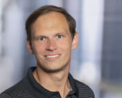 Eine Person mit kurzen Haaren und dunklem Hemd mit Kragen lächelt in die Kamera. Der sanft unscharfe Hintergrund lässt auf eine städtische Umgebung schließen, vielleicht in der Nähe der Hochschule Coburg, und verleiht der Szene einen Hauch akademischen Charme.