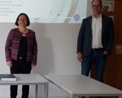 Eine Frau und ein Mann stehen in einem Raum neben einem Präsentationsbildschirm mit einer Uhr an der Hochschule Coburg. Sie lächeln beide, vor ihnen stehen leere Schreibtische. Der Mann trägt einen Blazer, die Frau eine gemusterte Jacke.