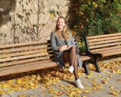 Eine Person sitzt auf einer Holzbank, umgeben von herbstlichem Laub in der Nähe der Hochschule Coburg. Sie trägt einen grauen Mantel und lächelt. Sie genießt das Sonnenlicht, das sanfte Schatten auf die Bank und die Steinmauer im Hintergrund wirft.