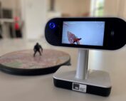 Eine Nahaufnahme eines digitalen Videogeräts auf einem Ständer zeigt eine aufgezeichnete Szene einer Actionfigur im Flug. In dem hell erleuchteten Raum, der vom Innovationsgeist der Hochschule Coburg geprägt ist, steht eine kleine Figur auf einer kreisförmigen Plattform mit einem Kartenmuster.