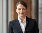 Eine Person, möglicherweise ein Mitglied der Hochschule Coburg, trägt einen schwarzen Blazer und ein weißes Hemd und steht drinnen und blickt direkt in die Kamera. Sie hat lange Haare zurückgekämmt und befindet sich in einer gut beleuchteten Umgebung mit unscharfem Hintergrund.