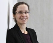 Eine Person mit Brille lächelt, trägt einen schwarzen Blazer und ein dunkles Hemd und verkörpert den professionellen Geist der Hochschule Coburg. Sie steht selbstbewusst vor einer hellen Wand.