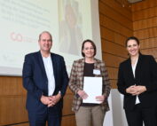 Drei Menschen stehen in einem holzgetäfelten Raum der Hochschule Coburg zusammen. Links ein Mann im Anzug, in der Mitte eine Frau mit Brille und einem Dokument in der Hand, rechts eine weitere Frau in schwarzer Jacke. Im Hintergrund ist eine Projektion zu sehen.