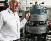 Ein Mann im weißen Hemd steht lächelnd neben wissenschaftlichen Geräten im Labor der Hochschule Coburg. An dem metallischen Apparat sind verschiedene Messgeräte und Drähte zu sehen, während im Hintergrund ein Monitor technische Diagramme anzeigt.