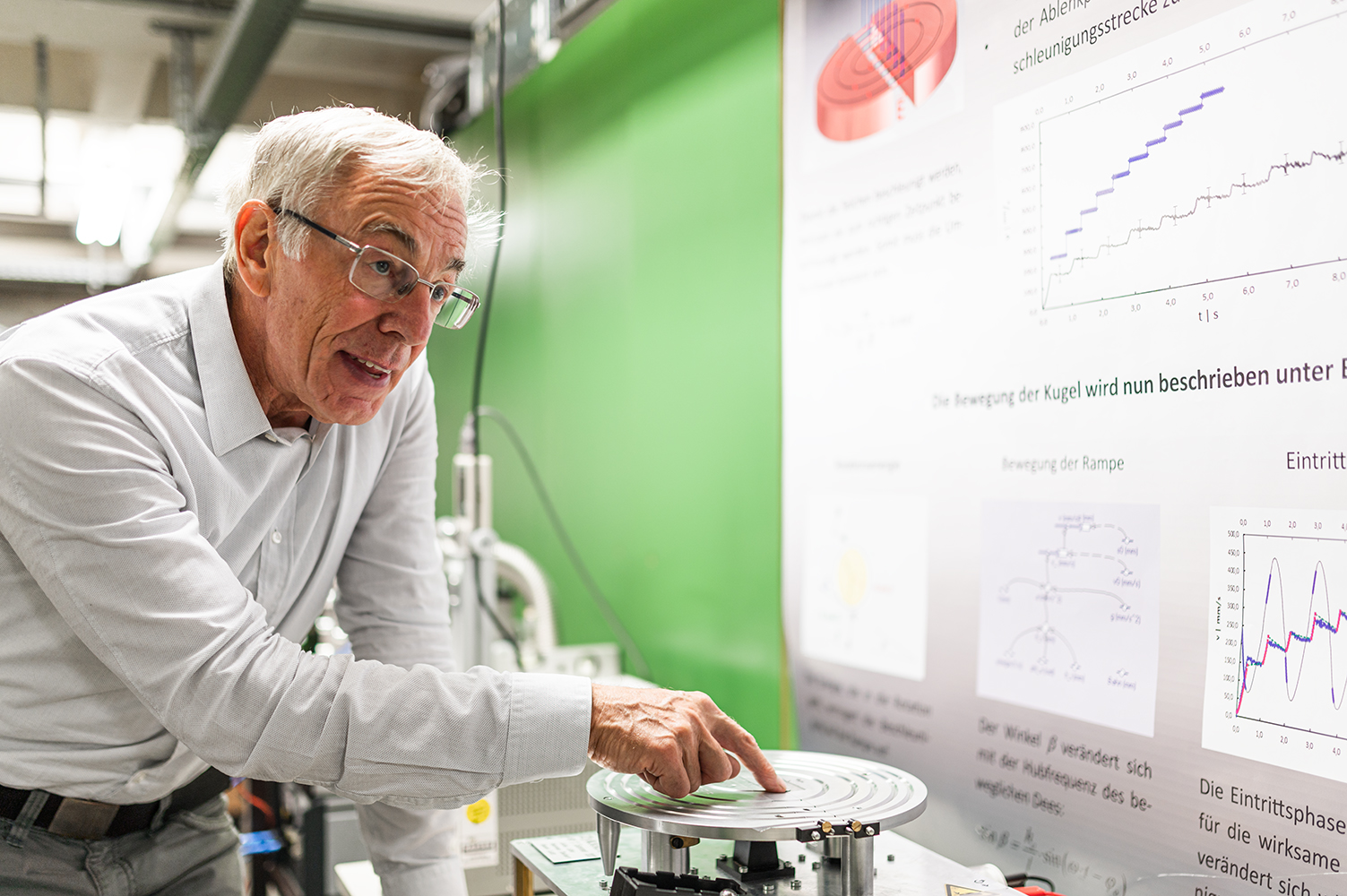 Ein älterer Mann mit Brille und weißem Hemd steht neben wissenschaftlichen Geräten der Hochschule Coburg und zeigt auf eine Scheibe. Hinter ihm schmücken Poster mit Grafiken und Diagrammen die grün-weißen Wände.