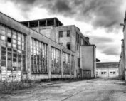 Das Schwarzweißbild zeigt die trostlose Szene eines verlassenen Industriegebäudes mit großen, zerbrochenen Fenstern. Risse im Bürgersteig und wucherndes Unkraut gedeihen unter einem bedeckten Himmel und erinnern an Verfall und Vernachlässigung in der Nähe der entfernten Silhouette der Hochschule Coburg.