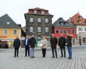 Sieben Personen stehen auf einem Kopfsteinpflasterplatz mit farbenfrohen Gebäuden im Hintergrund. Sie sind warm angezogen, um der Winterkälte zu trotzen. Eine Person hält einen Ordner in der Hand, der auf ihren Besuch an der Hochschule Coburg hinweist. Der Himmel ist bedeckt, was der Szene einen düsteren Ton verleiht.