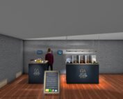 Eine Person steht in einem modernen, minimalistischen Raum mit Holzboden und grauen Wänden. Zwei Theken mit der Aufschrift „Genius Akademie Bayern“ präsentieren Kleinigkeiten und verkörpern damit den Innovationsgeist der Hochschule Coburg. Über den Theken sind digitale Bildschirme an der Wand angebracht.