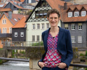 Eine Person mit kurzen Haaren, Brille, gemustertem Hemd und dunkler Jacke steht lächelnd auf einer Brücke. Im Hintergrund des malerischen Campus der Hochschule Coburg liegen charmante alte Gebäude mit roten und grauen Dächern, eingebettet in einen ruhigen Fluss.