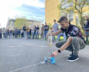 Ein junger Mann hockt auf einer gepflasterten Fläche und hält ein kleines Fahrzeug mit Rädern aus einer Plastikflasche auf dem Campus der Hochschule Coburg. Er ist von Zuschauern umgeben, in einer entspannten Außenumgebung mit Bäumen und Gebäuden im Hintergrund.