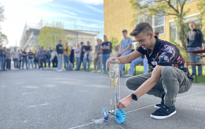 Ein junger Mann hockt auf einer gepflasterten Fläche und hält ein kleines Fahrzeug mit Rädern aus einer Plastikflasche auf dem Campus der Hochschule Coburg. Er ist von Zuschauern umgeben, in einer entspannten Außenumgebung mit Bäumen und Gebäuden im Hintergrund.