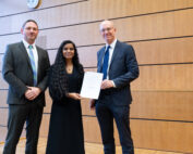 Drei Personen in formeller Kleidung stehen drinnen vor einer Holzwand in der Hochschule Coburg. Eine Frau in der Mitte hält ein Zertifikat, während zu beiden Seiten zwei Männer lächeln. Über ihnen ist eine Uhr zu sehen.