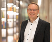 Ein Mann mit Brille, weißem Hemd und dunklem Blazer lächelt in die Kamera. Er steht in einem Innenbereich der Hochschule Coburg und zeigt vor einem unscharfen Hintergrund Bücherregale und moderne Beleuchtung.