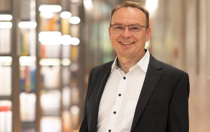 Ein Mann mit Brille, weißem Hemd und dunklem Blazer lächelt in die Kamera. Er steht in einem Innenbereich der Hochschule Coburg und zeigt vor einem unscharfen Hintergrund Bücherregale und moderne Beleuchtung.