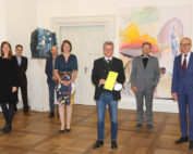 Eine Gruppe von sechs Personen steht in einem Raum mit Holzboden und Kunstwerken an den Wänden der Hochschule Coburg. Die Person in der Mitte hält ein gelbes Blatt Papier. Sie sind in formelle Kleidung gekleidet und der Raum wirkt hell und geräumig.