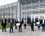 Sieben Personen in Business-Kleidung stehen vor einem modernen Gebäude der Hochschule Coburg mit Metallverkleidung. Drei Personen halten Zertifikate in der Hand, stehen etwas abseits, während andere lächeln. Die Szene lässt auf eine berufliche oder feierliche Veranstaltung schließen.