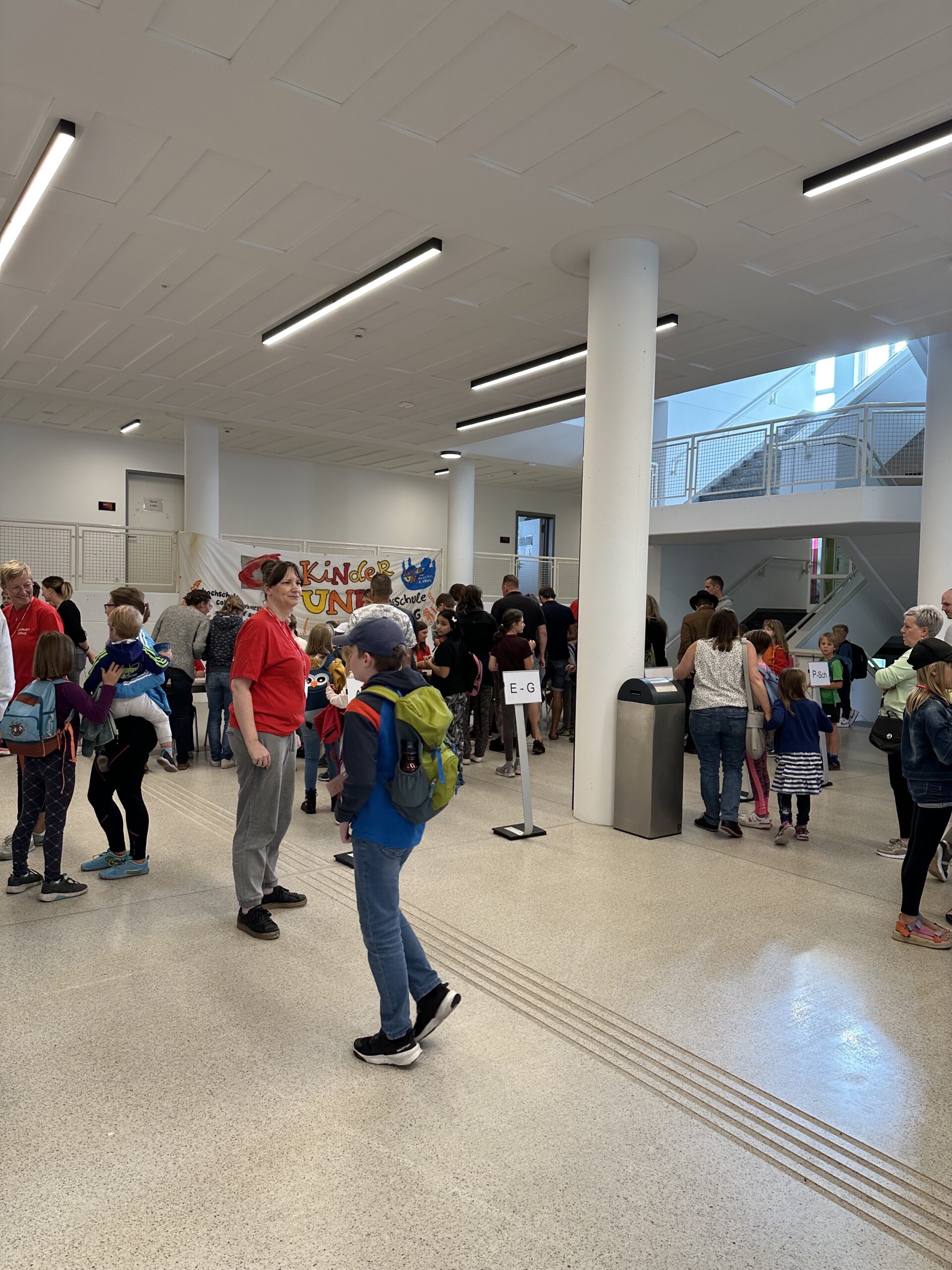 Eine Gruppe von Menschen, darunter Kinder und Erwachsene, versammelt sich im Foyer der Hochschule Coburg. Einige Kinder tragen Rucksäcke. Auf einem Banner steht „Make an Impact“. Im Hintergrund sind Schilder mit Buchstaben und eine Treppe zu sehen.