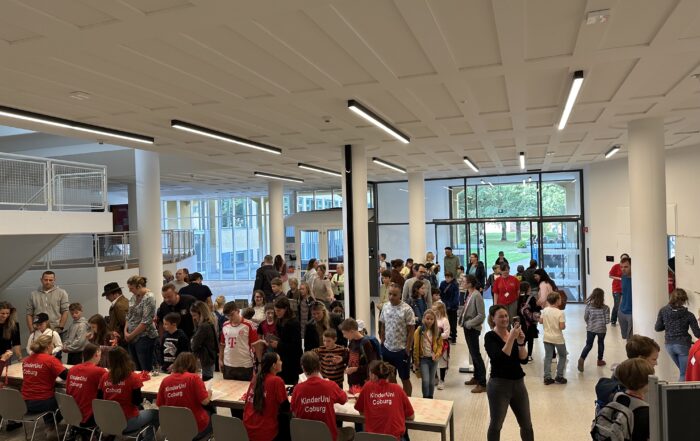 Bei der geschäftigen Veranstaltung in Innenräumen der Hochschule Coburg sind viele Menschen um Tische versammelt. Freiwillige in roten Hemden sitzen an einem Tisch und helfen den Teilnehmern. Der Raum verfügt über große Fenster und modernes Dekor, während sich Menschen unterschiedlichen Alters an lebhaften Gesprächen und Aktivitäten beteiligen.