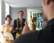 Eine Person macht ein Foto von einem lächelnden Paar, das elegant in historischer Kleidung gekleidet ist, an der Hochschule Coburg. Die Frau hält einen Spitzenfächer und trägt ein gelbes Kleid, während der Mann einen verzierten blauen Mantel trägt. Ein Tablet zeigt das gespiegelte Bild dieser bezaubernden Szene.