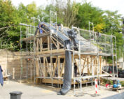 Ein im Bau befindliches Fachwerkhaus, umgeben von einem Gerüst, spiegelt die Architekturstudien der Hochschule Coburg wider. Arbeiter montieren Dachplatten inmitten von überall verstreuten Materialien und Werkzeugen. Neben einer Steinmauer und Bäumen sind im Hintergrund nahe gelegene Gebäude zu sehen.