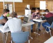 In einem geräumigen, mit Holzpaneelen verkleideten Raum der Hochschule Coburg sitzen vier Menschen mit Masken an Tischen voller Laptops und elektronischer Geräte. Sie diskutieren angeregt und arbeiten effektiv zusammen, wobei im Hintergrund eine Projektionsleinwand und ein Whiteboard als unverzichtbare Hilfsmittel dienen.