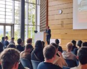 Eine Person steht an einem Rednerpult in einem modernen Hörsaal mit Holzwänden und großem Fenster an der Hochschule Coburg. Sie hält einen Vortrag vor einem sitzenden Publikum, während auf der Leinwand eine Folie angezeigt wird.