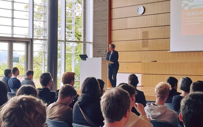 Eine Person steht an einem Rednerpult in einem modernen Hörsaal mit Holzwänden und großem Fenster an der Hochschule Coburg. Sie hält einen Vortrag vor einem sitzenden Publikum, während auf der Leinwand eine Folie angezeigt wird.
