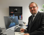 Ein Mann im Anzug steht an einem Schreibtisch mit einem Computermonitor, auf dem der Anmeldebildschirm der Hochschule Coburg angezeigt wird. Er hält einen Stift und ein Notizbuch in der Hand, im Hintergrund stehen Büromaterialien und Bücher auf Regalen. Auf dem Regal steht ein kleiner Globus, der für eine internationale Note sorgt.
