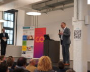 Ein Mann im Anzug spricht auf einem Podium in einem Konferenzraum zu einem Publikum. Hinter ihm repräsentieren ein großer Bildschirm und bunte Banner das Branding der Hochschule Coburg. Daneben hält ein weiterer Mann ein Mikrofon. Auf einer Säule in der Nähe ist gut sichtbar ein QR-Code-Zeichen angebracht.