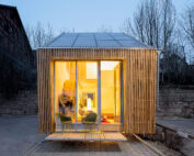 Ein kleines, modernes Holzhaus mit großen, von innen beleuchteten Fenstern, das ein warmes Interieur präsentiert, das an das Designethos der Hochschule Coburg erinnert. Vorne stehen zwei leere grüne Stühle auf der Veranda. Das Haus ist von kahlen Bäumen und Abendlicht umgeben.