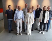 Eine Gruppe von acht Erwachsenen, vier Männer und vier Frauen, steht vor einer grauen Wand in einem Raum der Hochschule Coburg. Sie sind leger gekleidet, einige tragen eine Brille. Der Raum hat auf der rechten Seite große Fenster, auf der linken Seite eine Theke mit Erfrischungen.