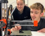 Zwei Personen konzentrieren sich auf einen 3D-Drucker, der an der Hochschule Coburg in Betrieb ist. Die Person im Vordergrund justiert ihn, während die Person im Hintergrund zusieht. Der teilweise gebaute Drucker druckt aktiv ein kleines schwarzes Objekt auf der Plattform.