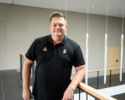 Eine Person im schwarzen Poloshirt lehnt an einem Holzgeländer in einem modernen Innenraum der Hochschule Coburg, umgeben von grauen Wänden und Hängelampen.