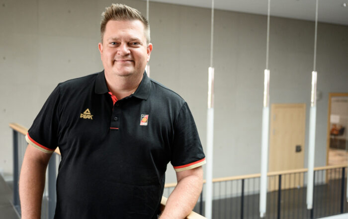 Eine Person im schwarzen Poloshirt lehnt an einem Holzgeländer in einem modernen Innenraum der Hochschule Coburg, umgeben von grauen Wänden und Hängelampen.