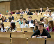 Im Hörsaal der Hochschule Coburg hört eine bunt gemischte Gruppe von Studierenden aufmerksam zu und macht sich Notizen. Der Raum ist mit Holzbänken und neutralen Farben gestaltet. Manche Studierende haben Laptops, andere schreiben in Notizbüchern.