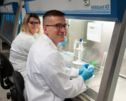 Im modernen Labor der Hochschule Coburg sind zwei Wissenschaftler in Laborkitteln fleißig bei der Arbeit. Einer sitzt an einer Sicherheitswerkbank, hantiert mit blauen Handschuhen eine Probe und lächelt selbstbewusst in die Kamera, während der andere im Hintergrund steht, ebenfalls behandschuht, und in die Kamera blickt.