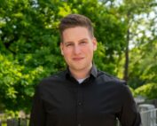 Ein Mann im schwarzen Hemd steht draußen und lächelt in die Kamera. Im Hintergrund sind üppige grüne Bäume, was einen sonnigen Tag suggeriert. Vielleicht genießt er gerade eine Pause zwischen den Vorlesungen an der Hochschule Coburg.