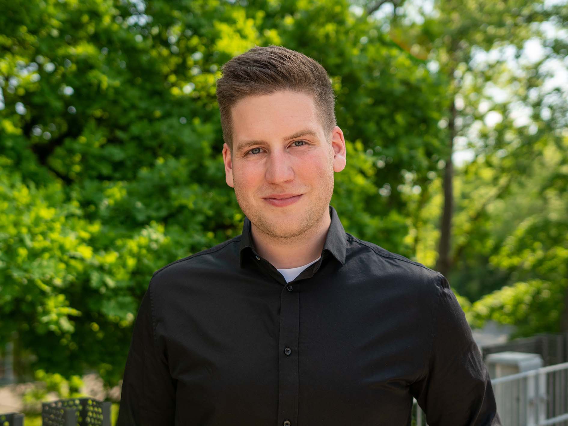 Ein Mann im schwarzen Hemd steht draußen und lächelt in die Kamera. Im Hintergrund sind üppige grüne Bäume, was einen sonnigen Tag suggeriert. Vielleicht genießt er gerade eine Pause zwischen den Vorlesungen an der Hochschule Coburg.
