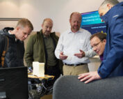 Eine Gruppe von fünf Männern in einem Raum der Hochschule Coburg sitzt um einen Schreibtisch herum und untersucht aufmerksam ein Gerät oder Modell. Ein Mann gestikuliert mit den Händen, während er spricht. Im Hintergrund ist auf einem Monitor der Schriftzug „Visualisierung von…“ zu sehen.