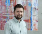 Eine Person mit kurzen Haaren und Bart trägt ein helles Hemd und steht vor einem abstrakten, farbenfrohen Gemälde mit roten, blauen und weißen Pinselstrichen an der Hochschule Coburg.