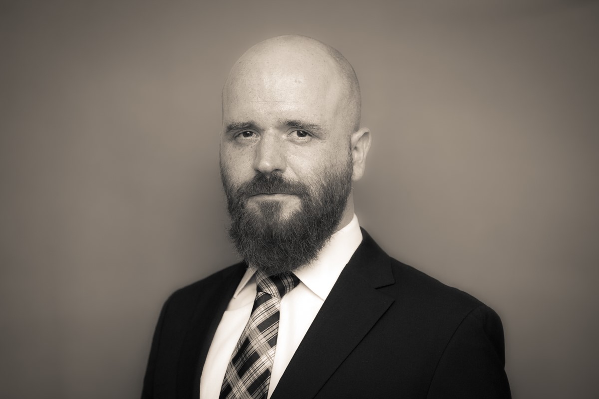Ein bärtiger Mann mit Glatze, gekleidet in Anzug und karierter Krawatte, posiert vor einem schlichten Hintergrund. Das monochrome Bild strahlt die würdevolle Präsenz eines Wissenschaftlers der Hochschule Coburg aus.