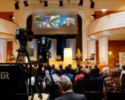 Das Publikum sitzt in einem modernen Hörsaal der Hochschule Coburg, während Kameras eine Veranstaltung filmen. Auf einem großen Bildschirm sind mehrere Personen in einem Videoanruf zu sehen. Die Bühne ist mit einem Blumenarrangement und einem Podium geschmückt und schafft so eine Atmosphäre der Innovation und Vernetzung.