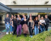 Eine Gruppe von fünfzehn Personen steht im Freien vor einem modernen Gebäude mit großen Fenstern der Hochschule Coburg. Sie tragen legere Kleidung und lächeln in die Kamera, umgeben von Grün und einem kleinen Wasserspiel im Vordergrund.