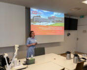 Eine Person steht in einem Raum und hält eine Präsentation an der Hochschule Coburg. Sie lächelt und steht neben einem großen Bildschirm, auf dem eine Folie mit Text und einem Bild eines Gebäudes läuft. Im Vordergrund steht ein Tisch mit einem Projektor und einigen Getränkeautomaten.