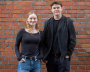 Zwei junge Erwachsene stehen vor einer Backsteinmauer an der Hochschule Coburg. Beide haben helle Haare und tragen legere dunkle Kleidung. Die Person links hat lange Haare, die Person rechts trägt eine Jacke. Beide strahlen ein entspanntes Auftreten aus.