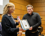 Bei einer offiziellen Veranstaltung an der Hochschule Coburg überreicht eine Frau einem Mann in einem holzgetäfelten Raum eine Urkunde. Im Hintergrund sind eine Projektionsleinwand und sitzende Zuschauer zu sehen. Beide Personen lächeln herzlich.