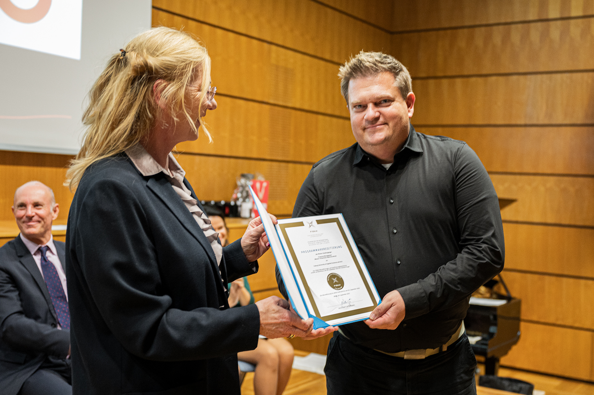 Bei einer offiziellen Veranstaltung an der Hochschule Coburg überreicht eine Frau einem Mann in einem holzgetäfelten Raum eine Urkunde. Im Hintergrund sind eine Projektionsleinwand und sitzende Zuschauer zu sehen. Beide Personen lächeln herzlich.