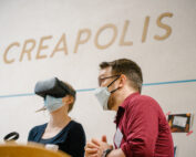 In einem Raum mit der Aufschrift „CREAPOLIS“ innerhalb der Hochschule Coburg sitzt eine Person mit VR-Headset neben einer Person mit Brille und Maske. Beide sind tief in ihre digitale Erkundung oder eine immersive Diskussion vertieft.
