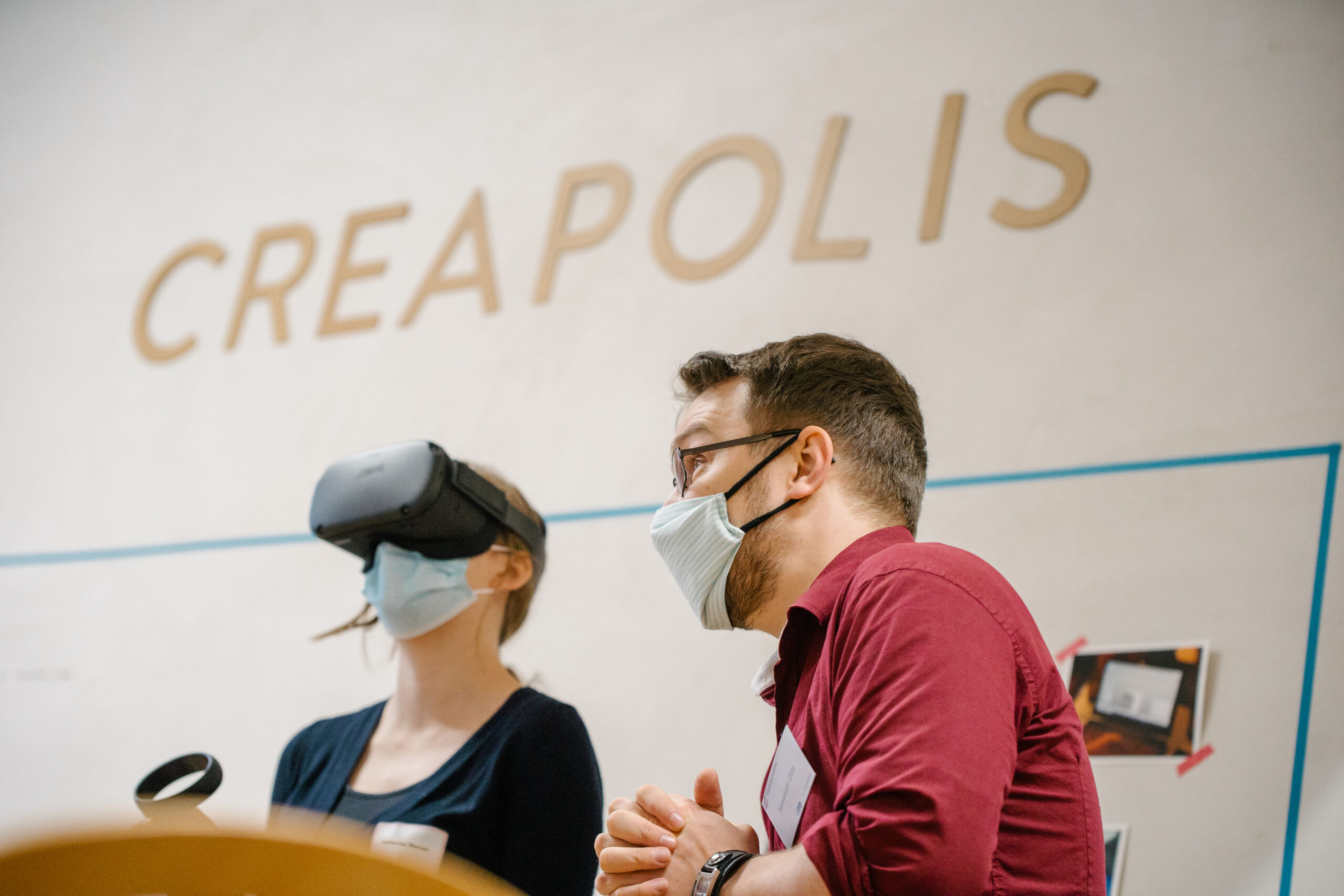 In einem Raum mit der Aufschrift „CREAPOLIS“ innerhalb der Hochschule Coburg sitzt eine Person mit VR-Headset neben einer Person mit Brille und Maske. Beide sind tief in ihre digitale Erkundung oder eine immersive Diskussion vertieft.