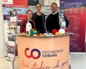 Drei Vertreter präsentieren hinter einem Promotion-Tisch Lehrmaterialien und Spielsachen der Hochschule Coburg. Im Hintergrund geben zwei große Banner Einblicke in die Welt der Bioanalytik.